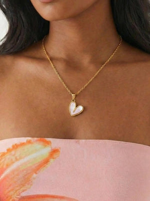 Woman wearing a gold necklace with a heart-shaped pendant, standing against a neutral background.