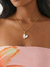 Woman wearing a gold necklace with a heart-shaped pendant, standing against a neutral background.